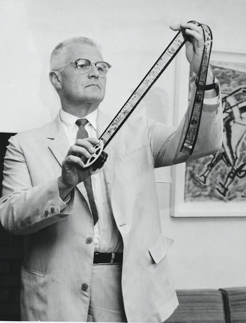 Eugene Power in a light suit, holding up and inspecting a long strip of microfilm from a reel